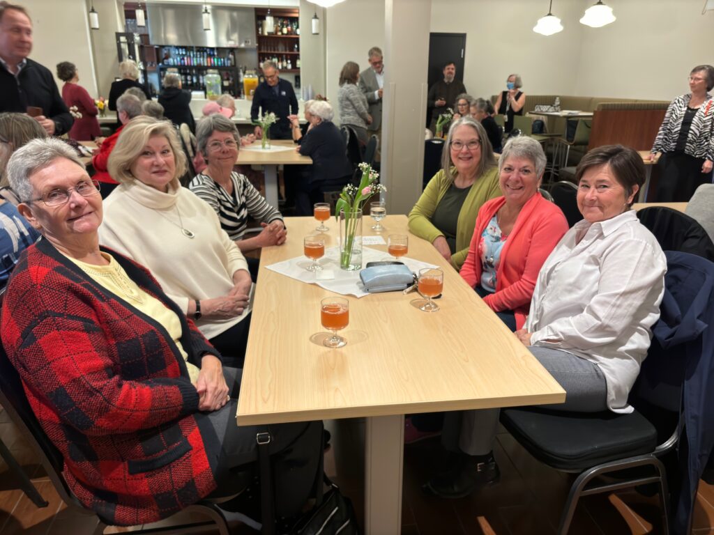 A group of smiling people sitting around a table in an event space with other people in the background